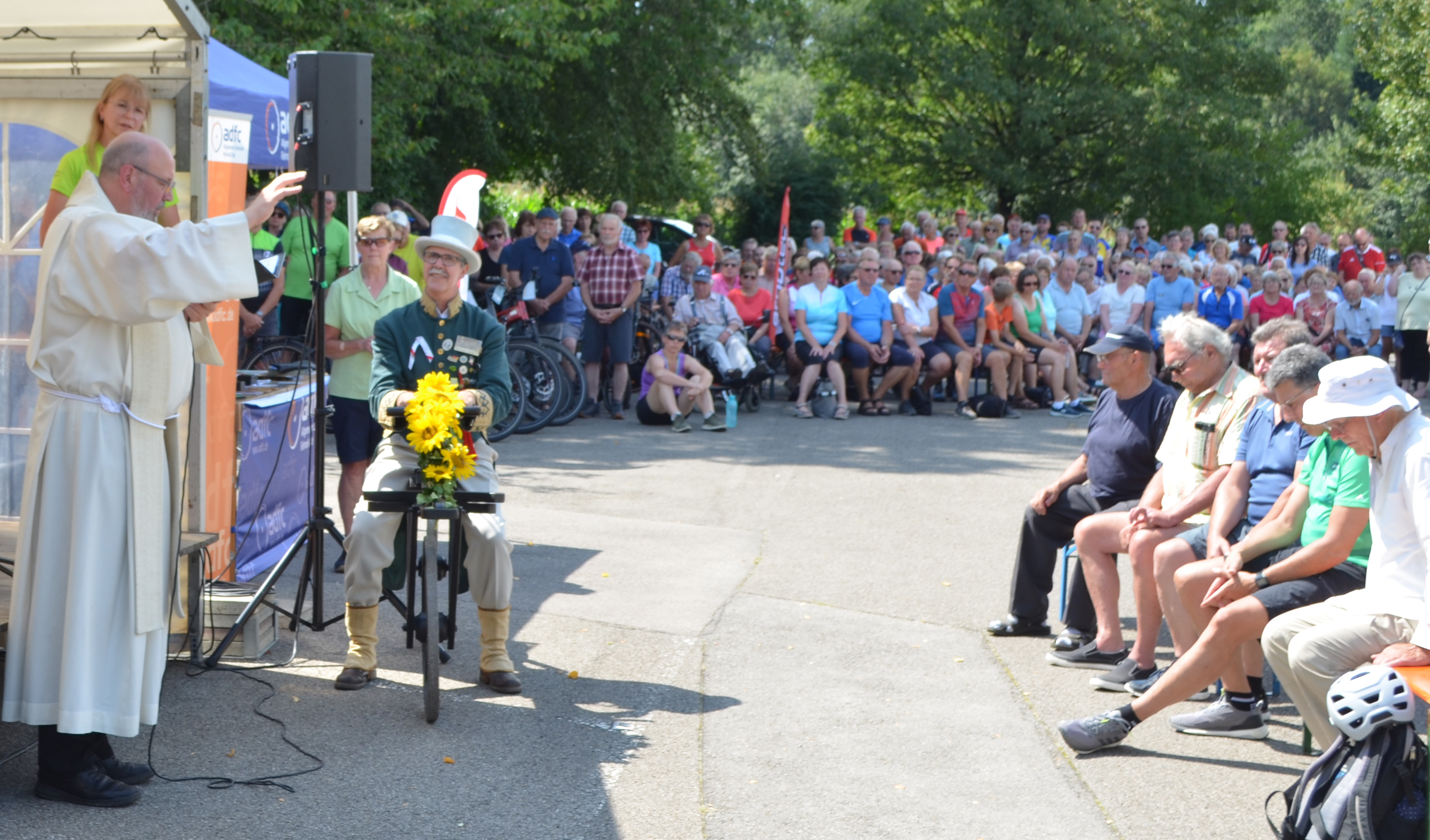 Links im Bild ein katholischer Pater, daneben sitzt ein Herr auf einem historischen Fahrrad, im Hintergrund sind viele Menschen zu sehen. 