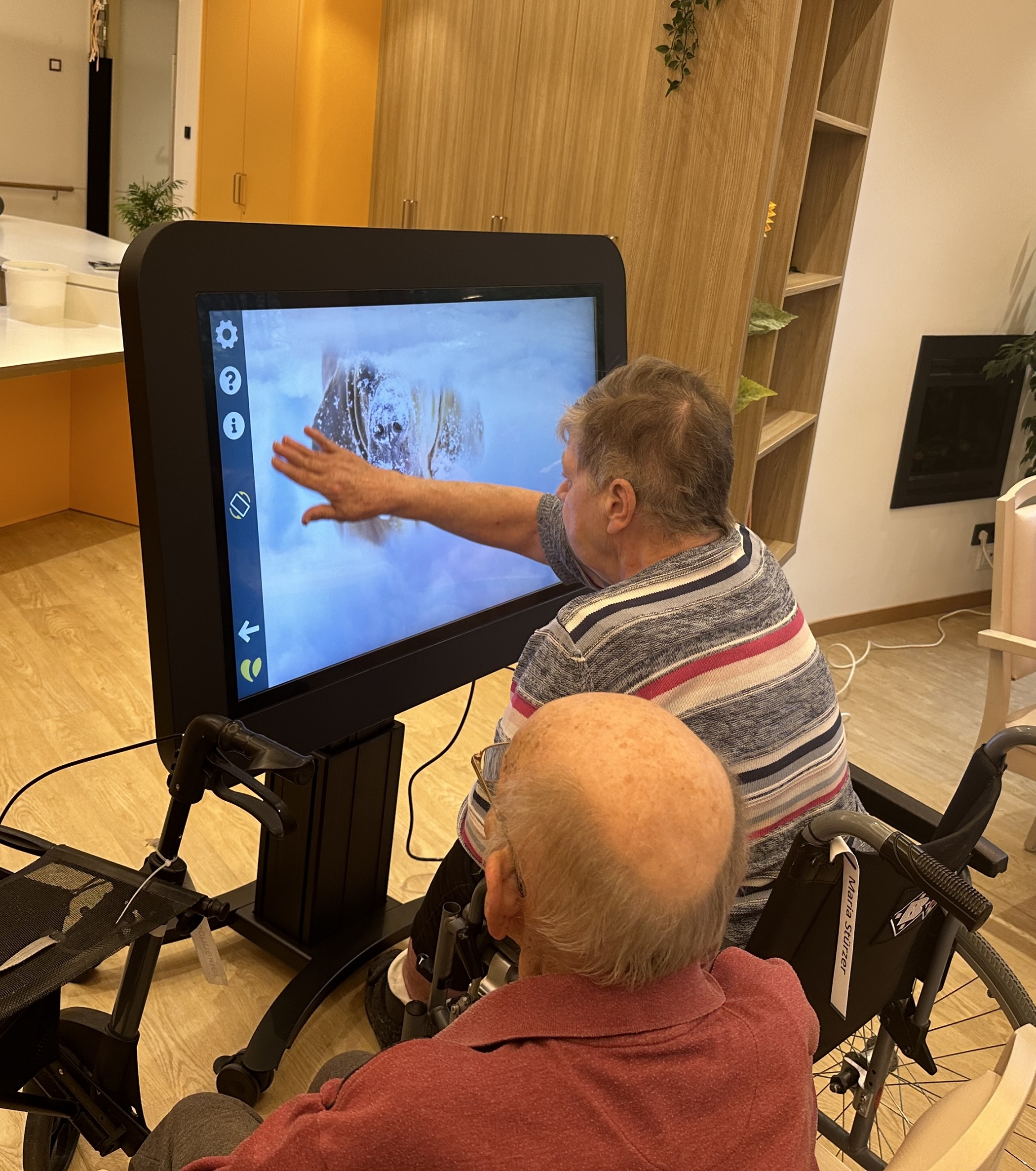 Zwei Senioren - eine Dame, ein Herr - sitzen vor einem großen Bildschirm. Die Dame berührt mit ihrer Hand den Bildschirm. 
