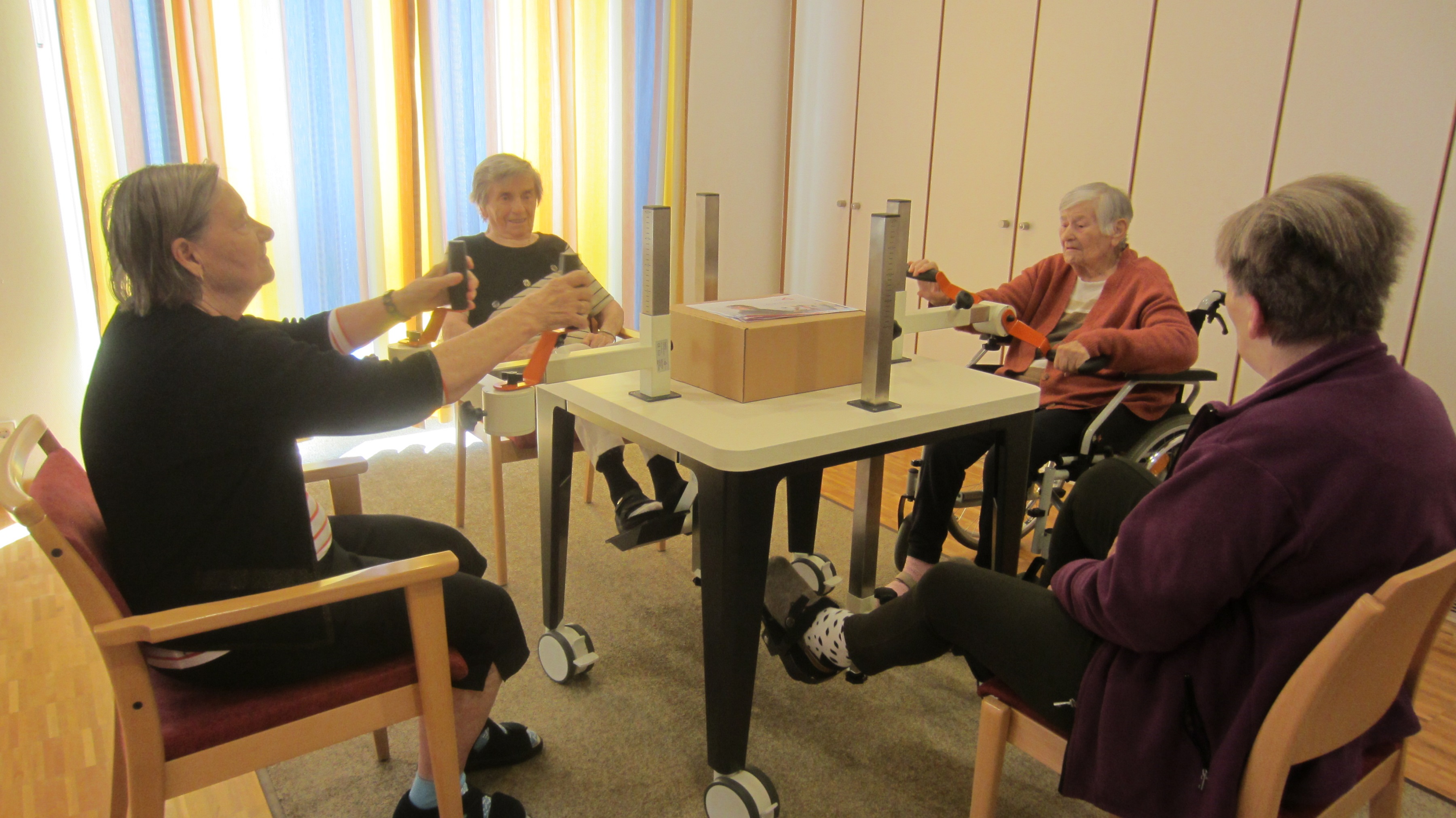 Vier ältere Damen sitzen um einen Tisch - eine tritt unter dem Tisch in die Pedale, die anderen betätigen Handkurbeln. 