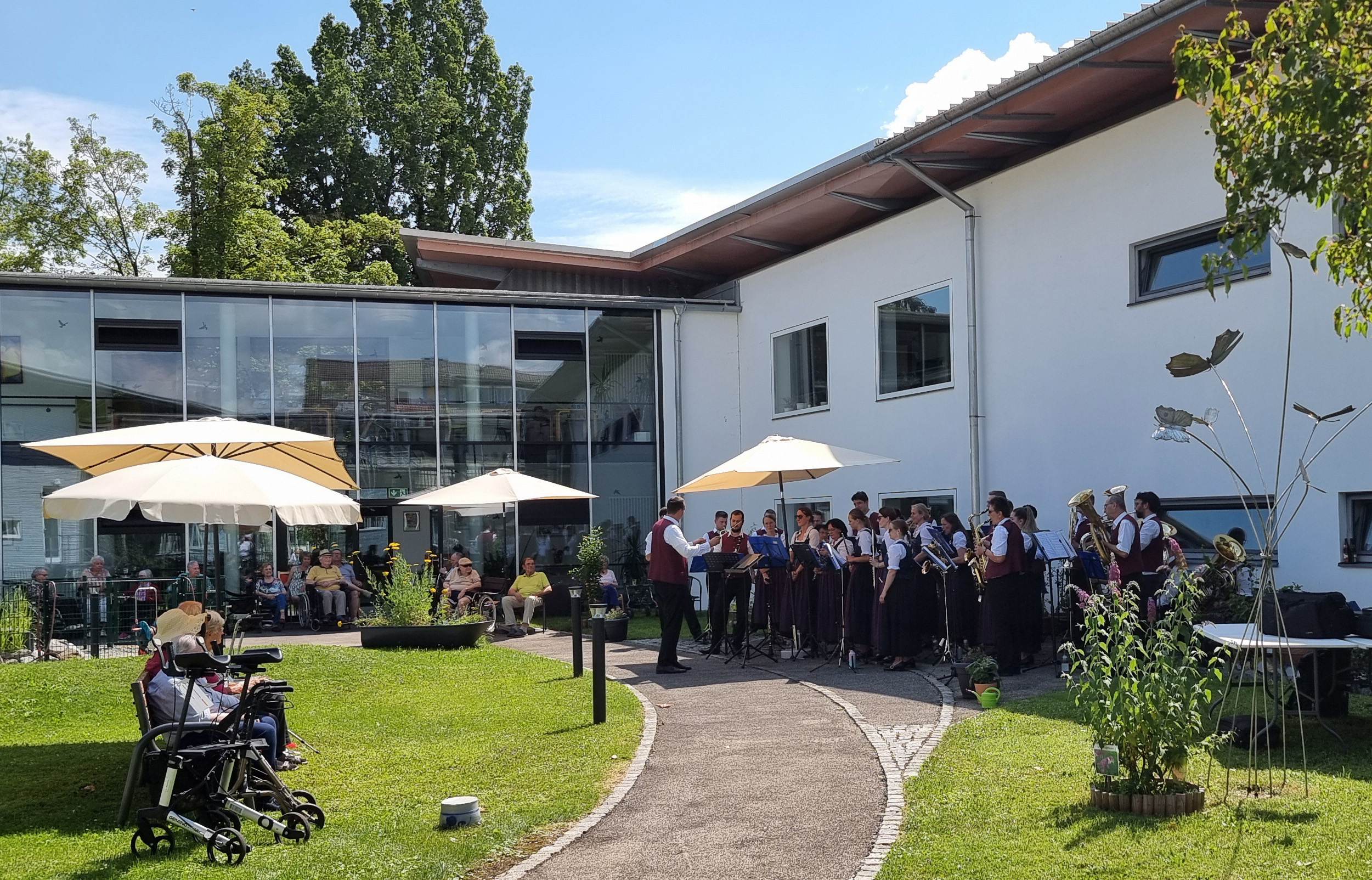 Die Stadtkapelle Bad Wörishofen spielt in einem Garten, darum sitzen die Seniorinnen und Senioren unter Sonnenschirmen. 