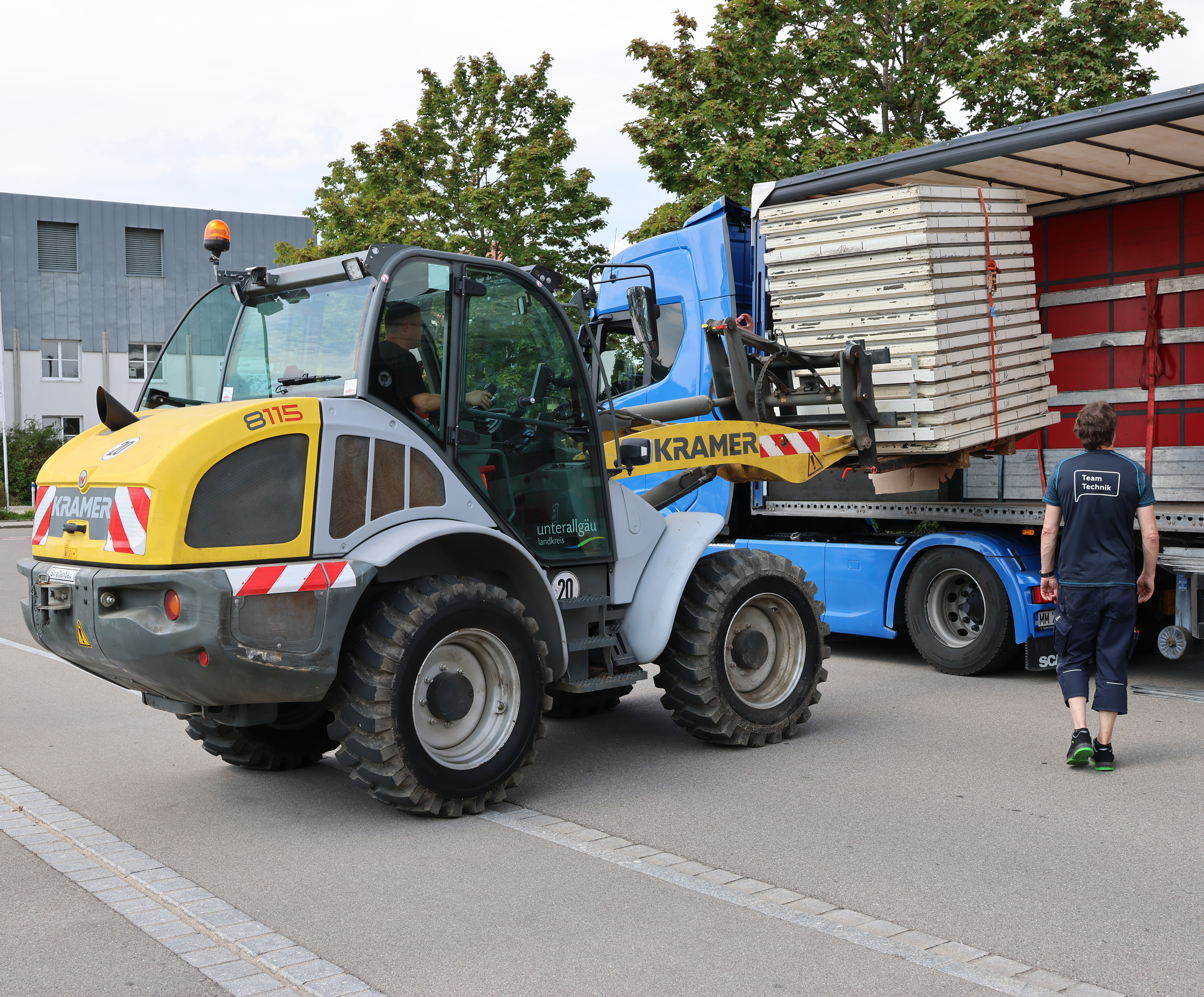 Ein Stapler steht vor einem großen Lastwagen und lädt eine Palette mit aufgestapelten Fenstern ein. 